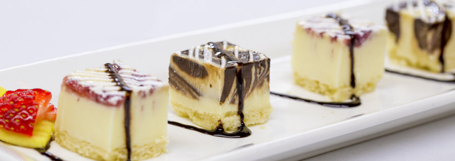 LUC Banquet / wedding hall catering deserts. A fruit garnished desert plate with a row of evenly spaced small delectable squares of various cheesecakes, all with a fine decorative zigzag line of chocolate sirup or liquor connecting it into one enticing desert.