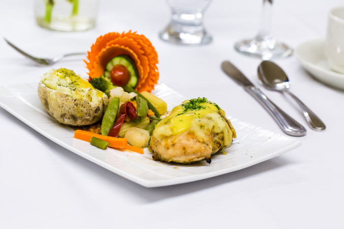 Banquet hall catering food, silverware and glasses. A garnished baked potato, sautéed or steamed veggies, stuffed chicken breast or dumpling covered in two kinds of melted cheese and sprinkled with parsley or cilantro. Decorative flower made of carrot, cucumber and grape tomato.