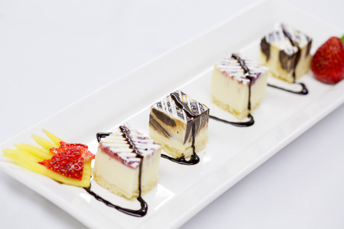 Banquet hall catering sweets. A fruit garnished desert plate with four small delectable squares of various cheesecakes, all covered with a fine decorative zigzag line of chocolate sirup or liquor.